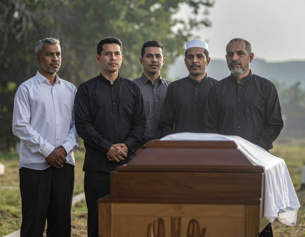 Un gruppo di uomini musulmani in preghiera comunitaria durante un funerale islamico, con una semplice bara di legno coperta da un sudario bianco in primo piano. L'immagine evoca la dignità, la semplicità e la solidarietà della comunità nel rito funebre islamico.